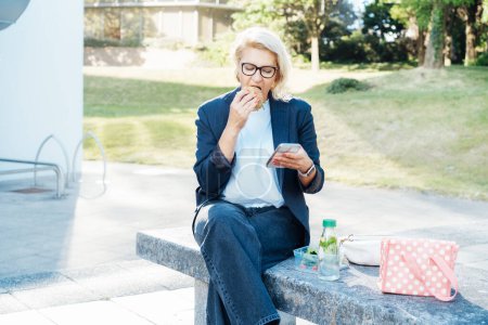 Femme d'affaires d'âge moyen mangeant un hamburger sandwich avec salade et utilisant le téléphone assis sur le banc dans le parc pendant la pause déjeuner. Travail à distance, entreprise, freelance, blogging, concept de médias sociaux.