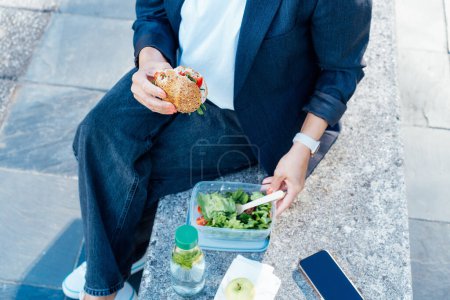 Téléchargez les photos : Vue de dessus gros plan femme d'affaires manger un sandwich et salade fraîche sur la pause déjeuner assis sur le banc au parc. Boîte à lunch régime équilibré pour la perte de poids. Habitudes alimentaires saines et bien-être. Concentration sélective - en image libre de droit