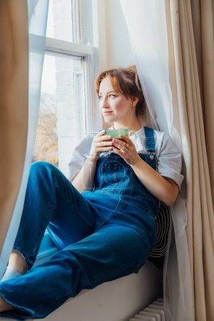 Time To Relax. Young woman looks out the window overlooking the city, sits on windowsill at cozy home holds cup of hot tea drink. Happy calm female taking break for mental health wellbeing. Vertical.