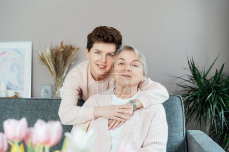 Senior 70s mother and adult neutral gender daughter hugging. Happy family enjoying weekend together in living room. Elderly woman spending quality time with daughter who visits her in retiring house