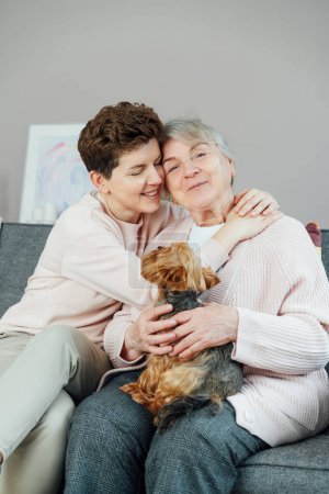 Portrait of happy senior 70s mother, adult neutral gender daughter and Yorkshire terriers dog hugging. Happy family enjoying weekend together in living room. Spending quality time in retiring house.