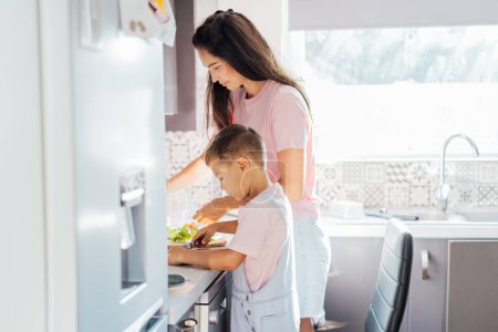 Loving young mother teaching her preschooler son prepare healthy food or salad at home kitchen, caring happy mom cooking together with kid on modern kitchen, boy involved in preparing food with mum.
