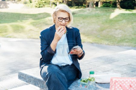Middle-aged business woman eating sandwich burger with salad and using phone sitting on bench in the park while having lunch break. Remote work,business, freelance,blogging,social media concept.