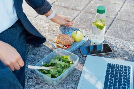 Téléchargez les photos : Vue de côté femme d'affaires ayant pause déjeuner, manger vite et en bonne santé devant un ordinateur portable à l'extérieur. Boîte à lunch équilibrée avec salade fraîche, sandwich, pomme, eau. Habitudes alimentaires saines et bien-être. - en image libre de droit