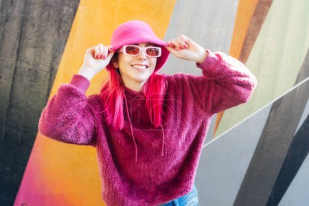 Hipster junge Frau mit pinkfarbenen Haaren und Sonnenbrille in magentaflauschigem Sweatshirt und Eimerhut posiert auf dem Wandhintergrund. Urbane Straßenmode. Einfarbige Optik. Gen Z, Millennials Selbstdarstellung