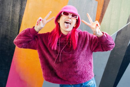 Emotional young hipster woman with pink hair and sunglasses in magenta fluffy sweatshirt and bucket hat making V sign by fingers on graffiti background. Urban street fashion. Gen Z self expression