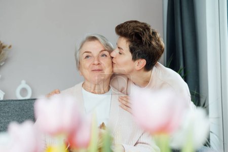 Eine erwachsene Frau küsst sich vor Vergnügen, eine ältere Mutter teilt einen zarten Moment mit verschwommenen Tulpen. Elternliebe, Fürsorge und Einheitskonzept. Muttertag. Glückliche Familie genießt Wochenende zusammen