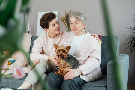 Senior 70s mother with dog pet and adult daughter share moment at home living room. Happy family enjoying weekend together. Elderly woman spending quality time with daughter in retiring house