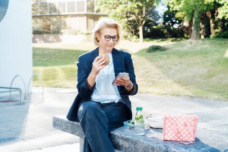 Mujer de mediana edad de negocios comiendo hamburguesa sándwich con ensalada y usando el teléfono sentado en el banco en el parque mientras almorza. Trabajo remoto, negocios, freelance, blogging, concepto de redes sociales.