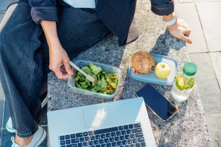 Top view business woman having lunch break, eating fast and healthy in front of laptop outdoors. Balanced diet lunchbox with fresh salad, sandwich, apple, water. Healthy eating habits and well-being