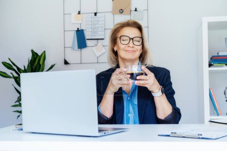 Successful relaxed dreamy middle aged business woman is resting at her work place, enjoying coffee break. Smiling confident middle-aged experienced senior female professional enjoying coffee drink.