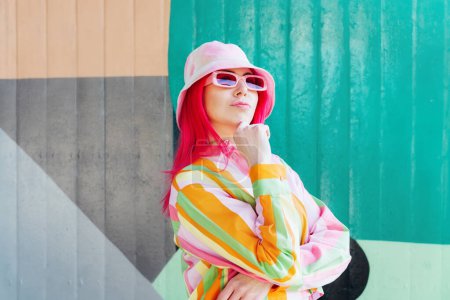 Urban street fashion look. Vanilla Girl. Kawaii vibes. Candy colors design. Bucket hat trends. Young woman with pink hair and sun glasses in multicolor strippled shirt on graffiti wall background