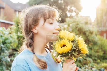 Dreaming young female farmer woman with closed eyes holding and sniffing a sunflowers bouquet on the green garden background in sunset light. Enjoy the moment. Rural, Cottage core lifestyle