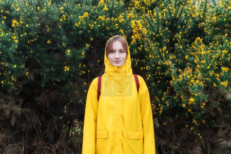 Téléchargez les photos : Jeune femme dans un imperméable jaune et capuche debout sur le fond de buisson fleuri avec des fleurs jaunes. Concept de voyage. Randonnée par tous les temps. Une fille en voyage. Détente, épanouissement personnel - en image libre de droit