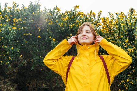 Smiling young woman in a yellow raincoat and hood with closed eyes enjoying the moment on walk with blooming bushes. Travel concept. Hiking in any weather. Girl on a journey. personal fulfillment.