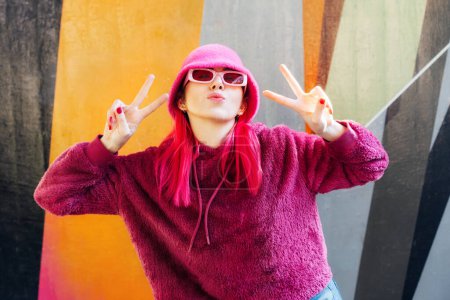 Emotionale junge Hipsterfrau mit pinkfarbenen Haaren und Sonnenbrille in magentaflauschigem Sweatshirt und Eimerhut, die mit den Fingern auf Graffiti-Hintergrund V-Zeichen setzt. Urbane Straßenmode. Gen Z Selbstdarstellung