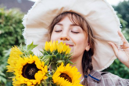 Nahaufnahme Träumende junge Bäuerin mit geschlossenen Augen, die einen Sonnenblumenstrauß auf dem grünen Gartenhintergrund hält und schnuppert. Genießen Sie den Moment. Ländlicher Lebensstil
