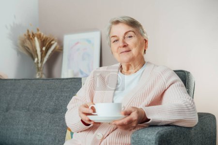 Lächelnde reife Frau, die eine Tasse Tee in der Hand hält, zu Hause entspannen, positive Seniorin, die zufrieden auf der Couch im modernen Wohnzimmer sitzt, fühlt sich wohl, ältere Generation, die das Leben genießt.