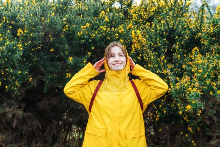 Foto de Mujer joven sonriente con un impermeable amarillo y capucha disfrutando de caminar sobre el fondo de un arbusto floreciente con flores. Concepto de viaje. Senderismo en cualquier clima. Una chica de viaje. Relajante, realización personal. - Imagen libre de derechos