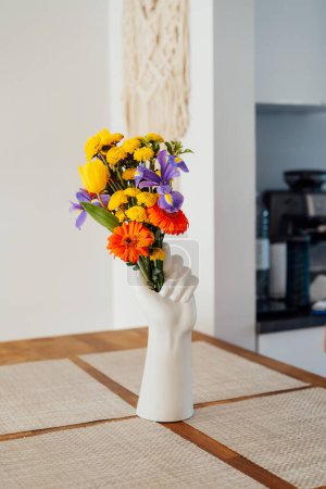 Téléchargez les photos : Table de comptoir de cuisine avec accent sur le vase en forme de main humaine avec bouquet de fleurs d'été multicolore divers avec fond flou de cuisine blanche confortable moderne. Détails de design d'intérieur - en image libre de droit