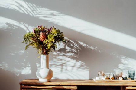 Téléchargez les photos : Intérieur scandinave avec bouquet exotique de fleurs de protéa dans un vase en céramique debout sur une armoire en bois sous la lumière du soleil et des ombres sur un mur gris blanc. Design minimaliste de décor à la maison - en image libre de droit