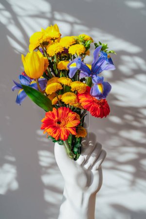 Téléchargez les photos : Vase en céramique fait en forme de main humaine avec bouquet de fleurs d'été multicolore sur table blanche sous la lumière du soleil et les ombres sur le mur gris. Détails de décoration intérieure tendance. Carte verticale - en image libre de droit