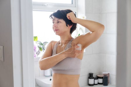 Young woman applying natural crystal alum deodorant in light modern bathroom. Eco friendly natural beauty and hygiene cosmetics. Zero waste, sustainable, alternative body care concept. Selective focus