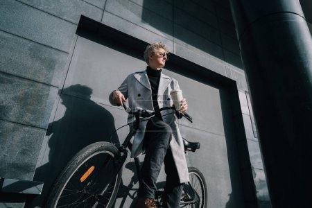 Stylish male in coat and sunglasses enjoying coffee out of reusable cup while sitting on retro bicycle near gray urban wall. Eco friendly transport for carbon footprint. Sustainable lifestyle