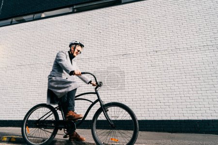 Stylish male in coat, sunglasses and protective helmet riding on retro bicycle near white brick urban wall. Neutral carbon footprint transportation. Green eco friendly mobility sustainable transport