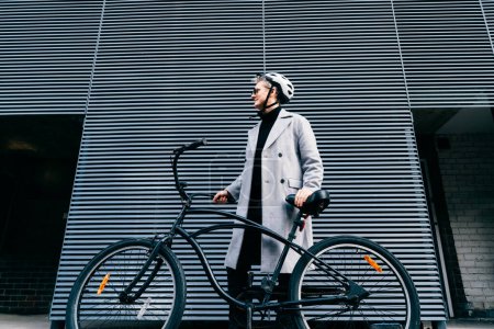 Téléchargez les photos : Homme élégant en manteau, lunettes de soleil et casque de protection debout avec vélo rétro près du mur urbain gris. Transport neutre de l'empreinte carbone. Mobilité écologique verte transport durable - en image libre de droit