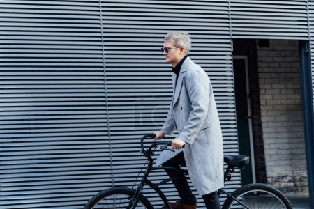 Téléchargez les photos : Homme élégant en manteau à carreaux et lunettes de soleil debout avec vélo rétro près du mur urbain gris dans une journée ensoleillée. Transport neutre de l'empreinte carbone. Mobilité écologique verte transport durable. - en image libre de droit