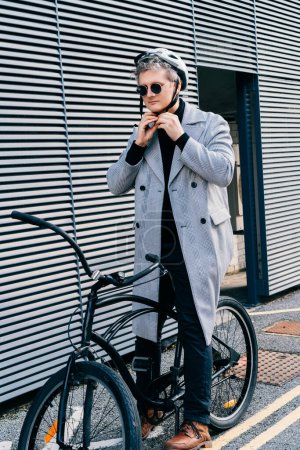 Stylish male in coat, sunglasses putting protective helmet on head before going to go ride by bicycle in city street. Neutral carbon footprint. Green eco friendly mobility sustainable transport