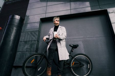 Téléchargez les photos : Homme élégant en manteau et lunettes de soleil profitant du café hors de tasse réutilisable tout en étant assis sur un vélo rétro près du mur urbain gris. Transport respectueux de l'environnement pour l'empreinte carbone. Mode de vie durable - en image libre de droit