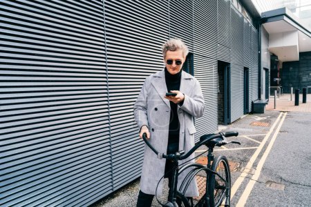 Stilvoller Mann in Mantel und Sonnenbrille mit Handy beim Spaziergang mit dem Retro-Fahrrad in der Stadt. Per Navigator-App. Neutraler CO2-Fußabdruck. Grüne umweltfreundliche Mobilität nachhaltiger Verkehr.