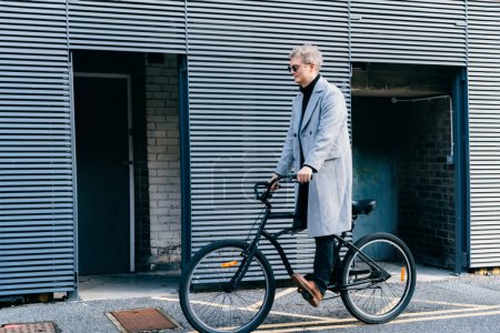 Stilvoller Mann in kariertem Mantel und Sonnenbrille steht mit Retro-Fahrrad an der grauen Stadtmauer bei sonnigem Tag. Neutraler Transport von CO2-Fußabdrücken. Grüne umweltfreundliche Mobilität nachhaltiger Verkehr.