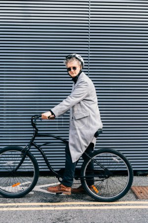 Stylish male in coat, sunglasses and protective helmet riding on retro bicycle near gray urban wall. Neutral carbon footprint transportation. Green eco friendly mobility sustainable transport