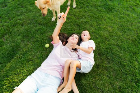 Téléchargez les photos : Enfant garçon mignon joyeux s'amusant avec sa mère, embrassant, riant sur l'herbe verte avec chien de compagnie dans le jardin au coucher du soleil. Activité de plein air estivale. Bonne enfance active, temps en famille. Concentration sélective. - en image libre de droit