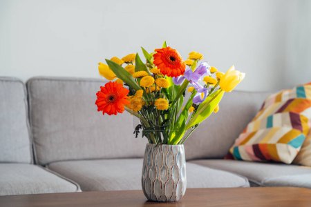 Téléchargez les photos : Vase en céramique avec bouquet de fleurs multicolore divers sur la table basse en bois de style vintage avec fond flou de salon moderne et confortable avec canapé gris. Design intérieur confortable à la maison - en image libre de droit