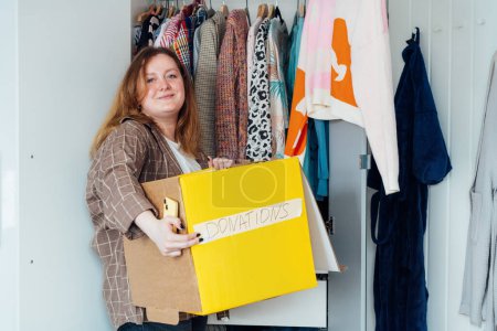 Smiling Woman with box of selected clothes from her wardrobe for donating to a Charity. Decluttering, Sorting clothes And Cleaning Up. Reuse, second-hand concept. Conscious consumer, sustainability.