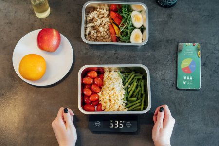 Healthy diet plan for weight loss, daily ready meal menu. Woman using meal tracker app on phone while weighing lunch box cooked in advance on kitchen scale. Balanced portion with dish. Pre-cooking.