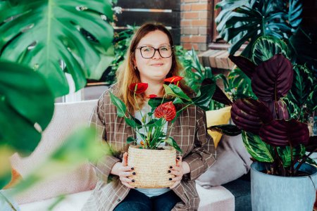 Portrait of Smiling Caucasian woman in casual clothes sitting on the sofa among home plants in her home garden conservatory. Home gardening hobby. Urban jungles. Biophilia lifestyle. Selective focus