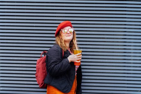 Téléchargez les photos : Femme souriante plus taille en forme de coeur lunettes de soleil et des vêtements lumineux boire du sucre aromatisé thé à bulles de tapioca près de mur urbain rayé. Joyeux hipster à la mode fille en surpoids. Mode de rue - en image libre de droit