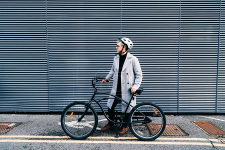 Stylish male in coat, sunglasses and protective helmet standing with retro bicycle near gray urban wall. Neutral carbon footprint transportation. Green eco friendly mobility sustainable transport.