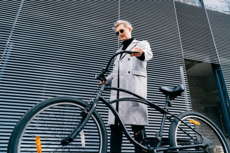 Stilvoller Mann in kariertem Mantel und Sonnenbrille, der bei sonnigem Wetter mit seinem Retro-Fahrrad in der Nähe der grauen Stadtmauer spaziert. Neutraler Transport von CO2-Fußabdrücken. Grüne umweltfreundliche Mobilität nachhaltiger Verkehr.