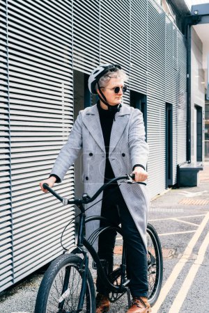 Stylish male in coat, sunglasses and protective helmet riding on retro bicycle near gray urban wall. Neutral carbon footprint transportation. Green eco friendly mobility sustainable transport