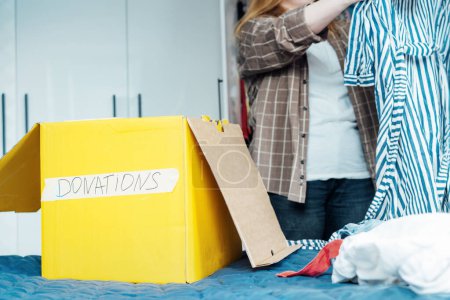 Boîte en carton jaune de vêtements sélectionnés pour faire un don à un magasin de charité debout sur le lit. Décluttage, tri des vêtements et nettoyage. Réutilisation, d'occasion. Consommateur conscient, durabilité.