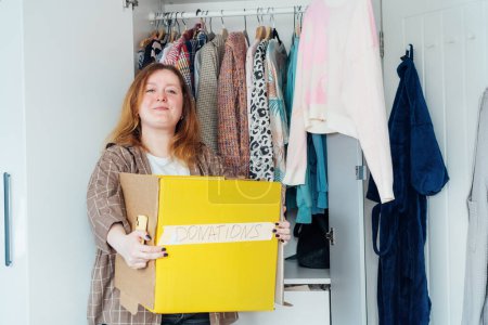 Smiling Woman with box of selected clothes from her wardrobe for donating to a Charity. Decluttering, Sorting clothes And Cleaning Up. Reuse, second-hand concept. Conscious consumer, sustainability.