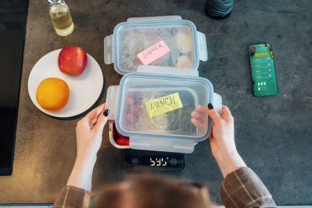 Gesunder Ernährungsplan zur Gewichtsabnahme, tägliches Fertigmenü. Close up Frau wiegt Lunchbox gekocht im Voraus, bereit, auf Küchenwaage zu essen. Ausgewogene Portion mit gesundem Gericht. Pre-cooking-Konzept