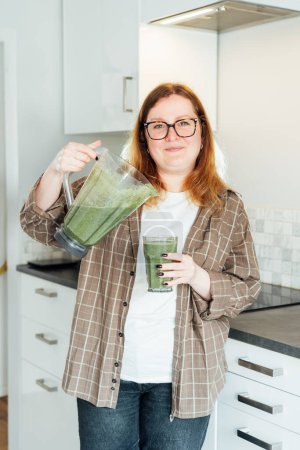 Smiling plus size young woman with just made glass of detox shake, green smoothie in the kitchen. Healthy dieting, eating, cooking, slimming. Natural weight loss program. Vegan, vegetarian diet