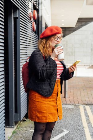 Young smiling plus size woman with backpack using mobile phone, drinking coffee outdoors on the street. Confident fashion hipster messaging, chatting, working remotely. Using navigator app for travel